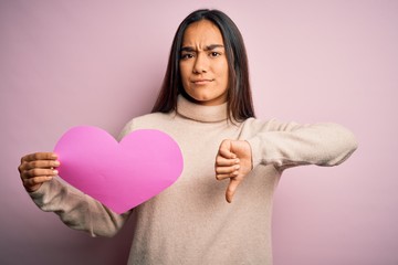 Canvas Print - Young beautiful asian woman holding pink heart standing over isolated background with angry face, negative sign showing dislike with thumbs down, rejection concept