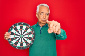 Poster - Middle age senior grey-haired man holding competition dartboard target over red background pointing with finger to the camera and to you, hand sign, positive and confident gesture from the front