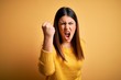 Young beautiful woman wearing casual sweater over yellow isolated background angry and mad raising fist frustrated and furious while shouting with anger. Rage and aggressive concept.