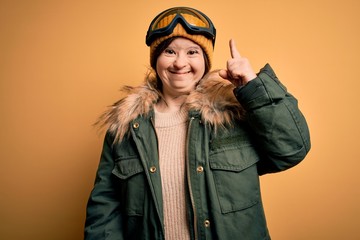 Sticker - Young down syndrome woman wearing ski coat and glasses for winter weather pointing finger up with successful idea. Exited and happy. Number one.