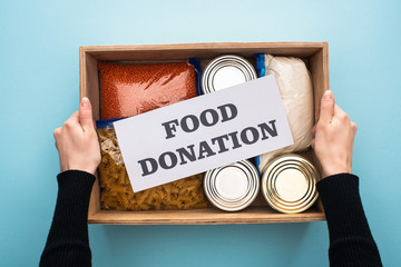 Wall Mural - cropped view of woman holding wooden box with cans and groats in zipper bags and card with food donation lettering on blue background
