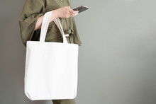 Blank White Tote Bag Canvas Fabric With Handle Mock Up Design. Close Up Of Woman Holding Eco Or Reusable Shopping Bag And Using Smartphone Near Grey Wall. No Plastic Bag And Ecology Concept.