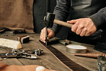 Working process of the leather belt in the leather workshop. Man holding hammer. Tanner in old tannery. Wooden table background. Close up man arm. Maintenance concept. Goods production.