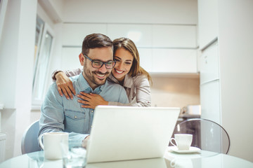 Wall Mural - Happy couple with laptop spending time together at home. Ordering online - Startup Business. Portrait of young excited couple hugging while working with laptop in cozy kitchen at home