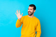 Young caucasian man over isolated blue background saluting with hand with happy expression