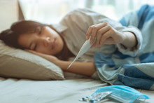 Asian Teen Infected With Covid-19 Flu Sick Lying In Bed Due To A Corona Virus Pandemic, Anxiously Measuring Check Body Temperature With Digital Thermometer.