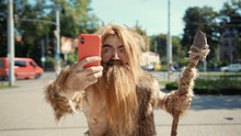 Portrait Of Wildman Hunter-gatherer Holding Modern Device Making Scary Fun Selfies Outdoor. Primeval Hipster Using Smartphone In Modern City Center.