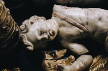 An Ancient Antique White Broken Marble Figure Of A Cupid With Broken Gilded Wings Lies On A Black Background Near The Tiara Of The Pope Close Up. The Cultural Heritage Of Classical Art.