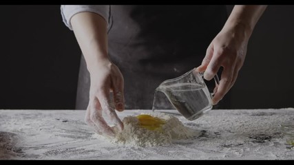 Wall Mural - Woman hands pour water into flour and eggs to knead homemade gough for preparation pasta on a black background. Slow motion, Full HD video, 240fps, 1080p.