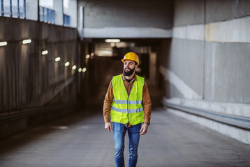 Wall Mural - Full length of hardworking attractive bearded worker in vest, with safety helmet on head exiting building in construction precess.