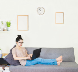 selective focus young beautiful asian woman wore glasses, brown t shirt and jean,sitting on gray sofa in living room ,work online lifestyle,relax at home with tea,home working laptop,work from home