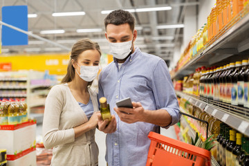 food shopping, consumerism and pandemic concept - happy couple wearing face protective medical masks for protection from virus disease with smartphone buying olive oil at grocery store or supermarket