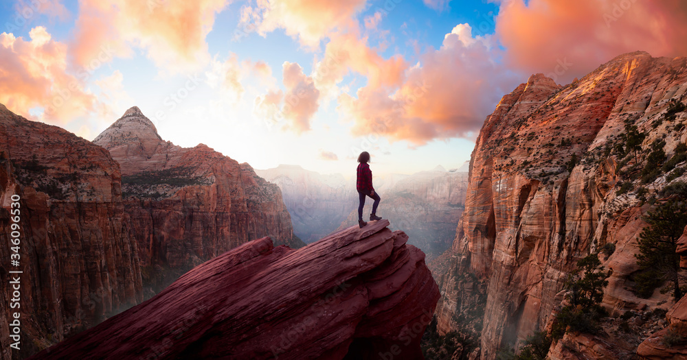 Adventurous Woman at the edge of a cliff is looking at a beautiful landscape view in the Canyon during a vibrant sunset. Taken in Zion National Park, Utah, United States. Sky Composite Panorama - obrazy, fototapety, plakaty 