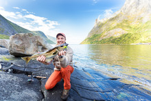 Happy Man With A Fish In His Hands. Fishing In Norway In The Fjord. Mountains On The Background.