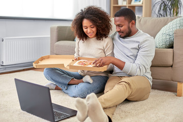 Wall Mural - leisure, people and food concept - happy african american couple with laptop computer eating takeaway pizza at home