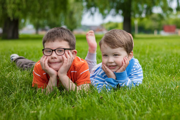 A portrait of two beautiful siblings who are lying on the green grass. One boy with glasses and an orange t-shirt. The second boy in a blue jacket. The boys are smiling.