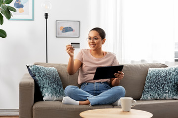 Wall Mural - internet banking, online shopping and technology concept - happy smiling african american woman sitting on sofa with tablet pc computer and credit card at home