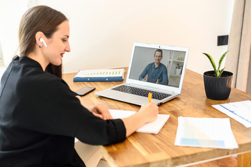 A male online tutor, teacher on the laptop screen, a young woman is watching webinar, online classes and writing a abstract, synopsis. Online learning concept