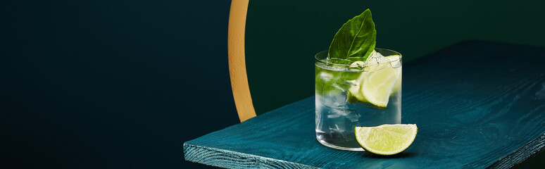 Wall Mural - Glass with refreshing drink with lime slice, ice and mint leaf on wooden surface on geometric blue and green background