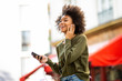 © mimagephotos - Side of happy young black woman listening to music with earphones and smart phone in city