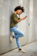 © mimagephotos - Full body young african woman with mobile phone and earphones leaning against wall