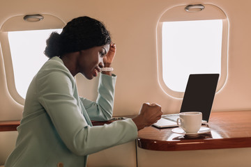 Wall Mural - smiling african american businesswoman near laptop with blank screen and coffee cup in private plane