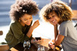 © mimagephotos - two young african american women sitting at outdoor cafe looking at cellphone