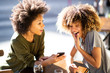 © mimagephotos - two cheerful young african american women sitting at outdoor cafe looking at cellphone