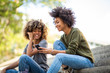 © mimagephotos - Side of two young african american women sitting on steps outside looking at mobile phone