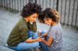 © mimagephotos - two young black women sitting on steps outside looking at mobile phone