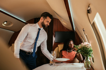 Wall Mural - selective focus of handsome air steward serving table near elegant african american woman in plane