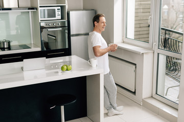 Wall Mural - man holding bowl with cornflakes and looking at window near apples, laptop and cup on table