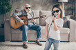 Photo of aged grandpa sitting comfy sofa playing guitar little pretty granddaughter hold mic singing acoustic song spend time together stay home quarantine modern living room indoors