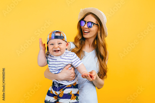 Fotomural Happy beautiful mother in a hat and sunglasses, holds a little child, on an isolated yellow background
