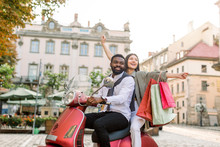 Cheerful Glamorous Young Multiethnic Couple Riding Red Vintage Scooter In The Street, African Man Wears White Shirt And Pants And Caucasian Woman Holds Colorful Shopping Bags With Travel Souvenirs