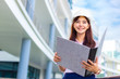 © kudosstudio - Happy young attractive female Asian engineer wearing hard hat holding her large size blueprint folder while standing outside of office building