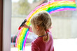 © Irina Schmidt - Adoralbe little toddler girl with rainbow painted with colorful window color during pandemic coronavirus quarantine. Child painting rainbows around the world with the words Let's all be well.