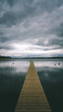 Fototapeta Na drzwi - Wooden jetty on a lake with dark blue clouds and sky reflection on water. Location is Vastersjon, Angelholm, Scania, Sweden.