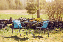 Small Table Set In Rural Home Garden With Chairs And Table For Two, Freshly Baked Pastries On Table, Sunny Spring Morning. Stacked Stone Fence Wall On Background. Blurred Field And Forest On.