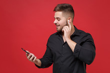 Smiling Young Bearded Guy In Classic Black Shirt Posing Isolated On Bright Red Wall Background Studio Portrait. People Emotions Lifestyle Concept. Mock Up Copy Space. Using Air Pods And Mobile Phone.