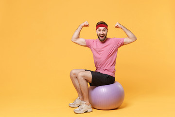 Wall Mural - Excited young bearded fitness sporty guy sportsman in headband t-shirt in home gym isolated on yellow background. Workout sport motivation lifestyle concept. Sit on fitball, showing biceps, muscles.