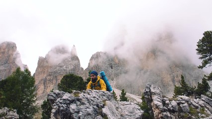 Wall Mural - young man hiker hiking up stone mountain with cloudy sky and fog. yellow jacket, backpack, black bea