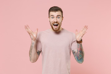 Surprised young bearded tattooed man guy in pastel casual t-shirt posing isolated on pink background studio portrait. People lifestyle concept. Mock up copy space. Keeping mouth open, spreading hands.