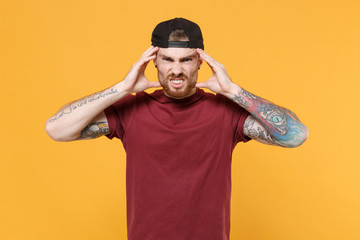 Displeased tired young bearded tattooed man guy in casual t-shirt, black cap posing isolated on yellow wall background. People lifestyle concept. Mock up copy space. Put hands on head having headache.