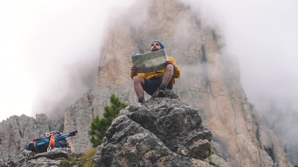 Wall Mural - young man hiker sitting on stone mountain reading map, with cloudy sky and fog. yellow jacket, backp