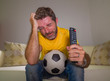 frustrated football fan in intense emotion - home portrait of young dejected and sad man watching soccer game on television at living room couch his team losing