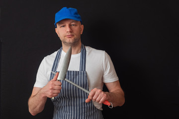 Portrait of a professional butcher in black and white apron, white t shirt and blue baseball hat. Sharpening his knives on a steel bar. Meat industry concept.