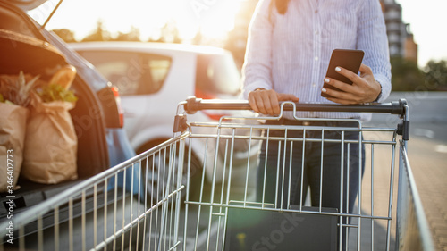 Close up woman hand using smart phone and holding trolley bar for shopping on sunny background at outside mall concept. Closeup of woman with shopping cart and shopping list in smartphone in hand