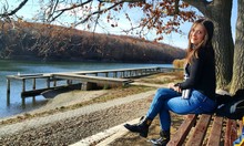 Side View Full Length Portrait Of Beautiful Woman Sitting By Lake On Bench