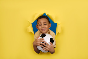 Smiling African-American boy soccer player and soccer fan in yellow Polo holds soccer ball in his hands against yellow torn paper wall background with space for text.